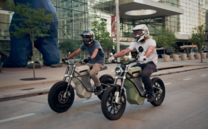 Two men riding District E Moto bikes through a city